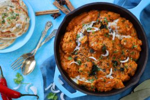Stock photo showing elevated surface view of turquoise blue pan of chicken tikka curry sauce. Butter chicken breast chunks sprinkled with red onion and mustard seeds, served with lachha paratha (layered flatbread).